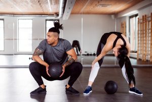 In a gym room, a young man squats down holding a medicine ball, looking over to the left, while a young woman beside him stands with her feet apart, bending forward with a medicine ball on the ground below her.