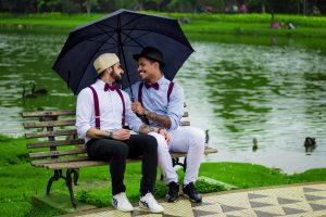 two young men sit on a bench near a pond, looking at each other smiling. One of them is holding an umbrella over their heads, and they are both wearing burgundy bow ties and suspenders.