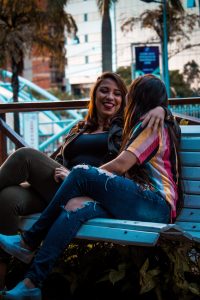 Two women sit on a bench in Brazil, looking at each other and laughing. The further woman has her arm around the other and you can see part of the city blurred out behind them.