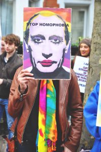 a protestor holds an artistic poster in front of their face of Russian President, Vladimir Putin, wearing makeup. Text scrolls across his head reading “stop homophobia” with a rainbow in the background.