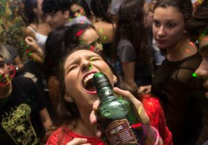 in the foreground you see a young woman tilting her head back with her mouth open, and another hand tipping a bottle of alcohol toward her as if she is going to drink it. Other young adults are partying around her.
