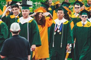 University grads smile and walk across campus wearing gold and green robes.