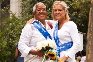image depicts an interracial lesbian couple who were able to marry in the USA, both wearing white, with a blue sash reading “Married” and holding flowers.