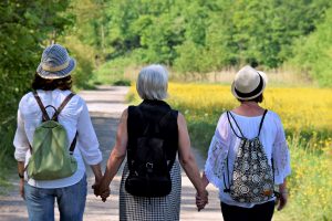 Three middle-aged women stand with their backs facing the camera, holding hands, appearing to be going for a walk.