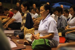 A middle-aged woman in the foreground sits with legs crossed, hands in lap, and eyes closed, in a state of meditation. There are other people in similar positions slightly blurred behind her, and you can see Buddhist Monks sitting in the background.