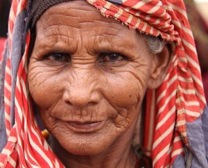Older woman from Somalia, with pronounced wrinkles, wearing a red-white striped head wrap stares into the camera with a smirk