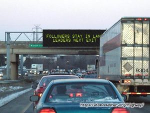 Cars driving on road with overhead sign saying Followers stay in lane, Leaders next exit.