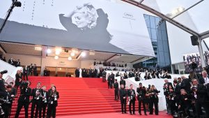 Personne en robe de soirée longue noire et dorée posant sur le tapis rouge devant l’entrée du Palais des Festivals à Cannes, avec une grande affiche du festival en arrière-plan.