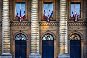 Façade d’un bâtiment officiel français avec trois grandes colonnes, des drapeaux tricolores et les mots « Liberté », « Égalité », « Fraternité » gravés au-dessus des portes.