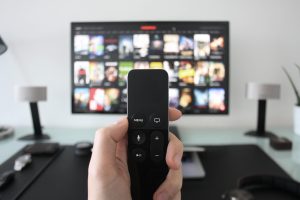 Main tenant une télécommande devant un écran de télévision affichant des vignettes de films ou séries, dans un salon moderne.