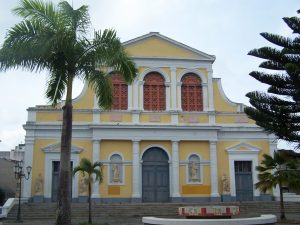 Façade de la cathédrale Saint-Pierre-et-Saint-Paul de Pointe-à-Pitre, en Guadeloupe : bâtiment jaune et blanc de style néoclassique, avec trois grandes portes grises, des colonnes blanches, des statues religieuses dans des niches, et des fenêtres à motifs rouges au niveau supérieur. Deux palmiers encadrent l’entrée, et un escalier mène à la place devant l’édifice.