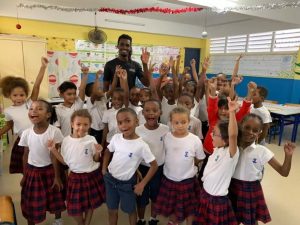 Groupe d’enfants en uniforme scolaire blanc et jupe ou short à carreaux rouges, rassemblés dans une salle de classe colorée autour d’un adulte debout, tous levant les bras avec enthousiasme. Des décorations festives et des affiches éducatives ornent les murs.