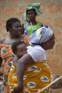 Trois femmes africaines en tenue traditionnelle colorée, l’une portant un enfant dans le dos avec un pagne jaune à motifs, se tiennent devant un mur en briques.