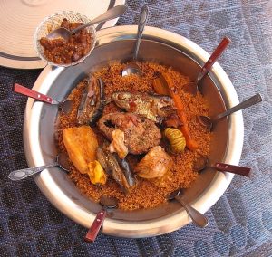 Grand plat rond de thiéboudiène (riz wolof), garni de poisson, légumes variés (carotte, chou, aubergine, manioc) et piment entier, entouré de plusieurs cuillères. À côté, un petit bol de sauce épicée avec une cuillère.