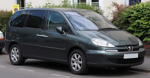 Monospace Peugeot 807 gris foncé, cinq portes, garé le long d’un trottoir devant des buissons et une maison.