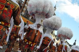 Personnes en costumes traditionnels colorés défilent dans une rue pavée bordée de bâtiments anciens lors d’un carnaval folklorique.