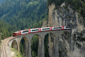 Un train rouge et blanc traverse un pont en arc de pierre à plusieurs arches, suspendu au-dessus d’une vallée boisée
