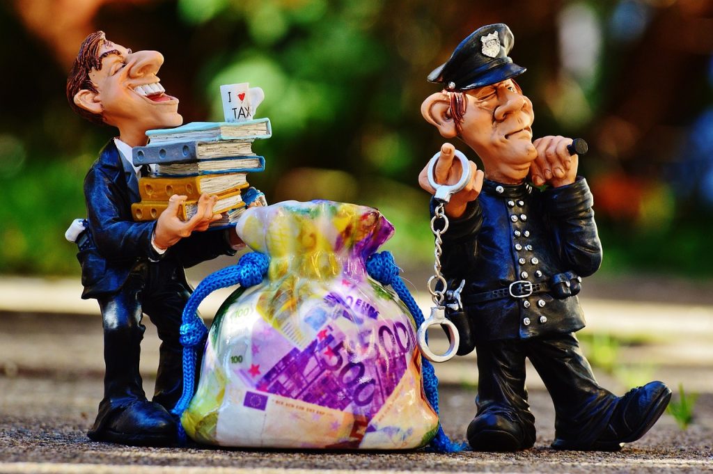 Figurines of police officer walking with handcuffs with a business manager holding a stack of books with a money bag behind him; background is out of focus pavement, sidewalk, and trees