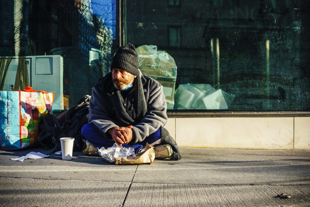 Man in a jacket sitting crossed legged on a street corner with a paper cup before him.