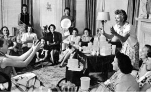 A black and white photo of a group of women sitting in a living room. Near the front of the room, a smiling woman tosses a tupperware container to a woman slightly out of frame with outstretched arms.