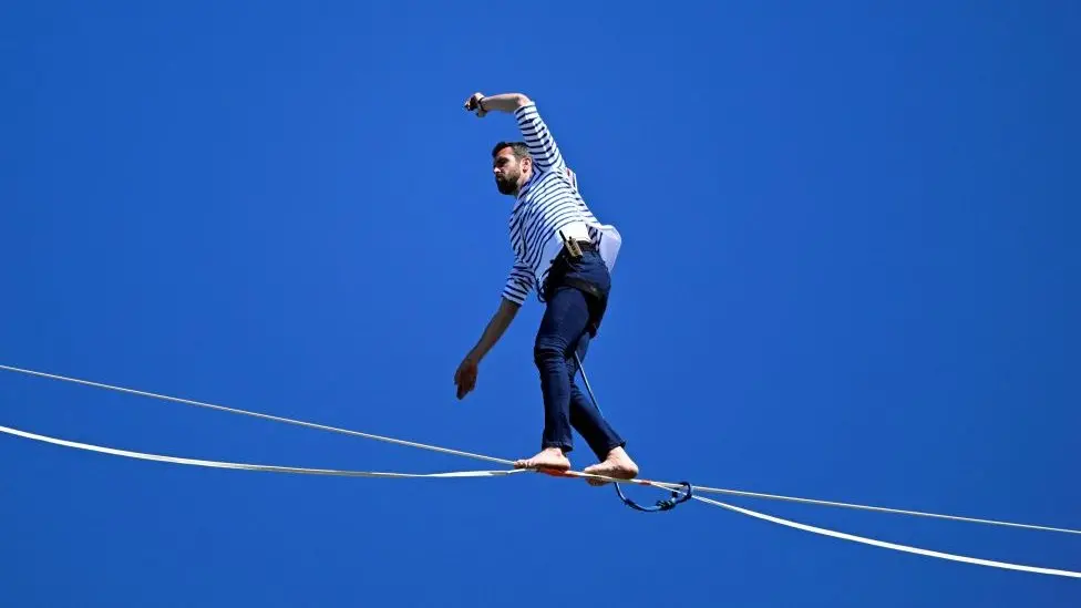 A man in a striped shirt and dark pants tightrope walking with his arms above his head on the background of a perfect blue sky.