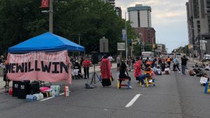 This is a picture of protesters standing in front of Hamilton City Hall. There is a blue tent, where protesters are gathered to voice their concerns about the Police Liaison Program in 2020.