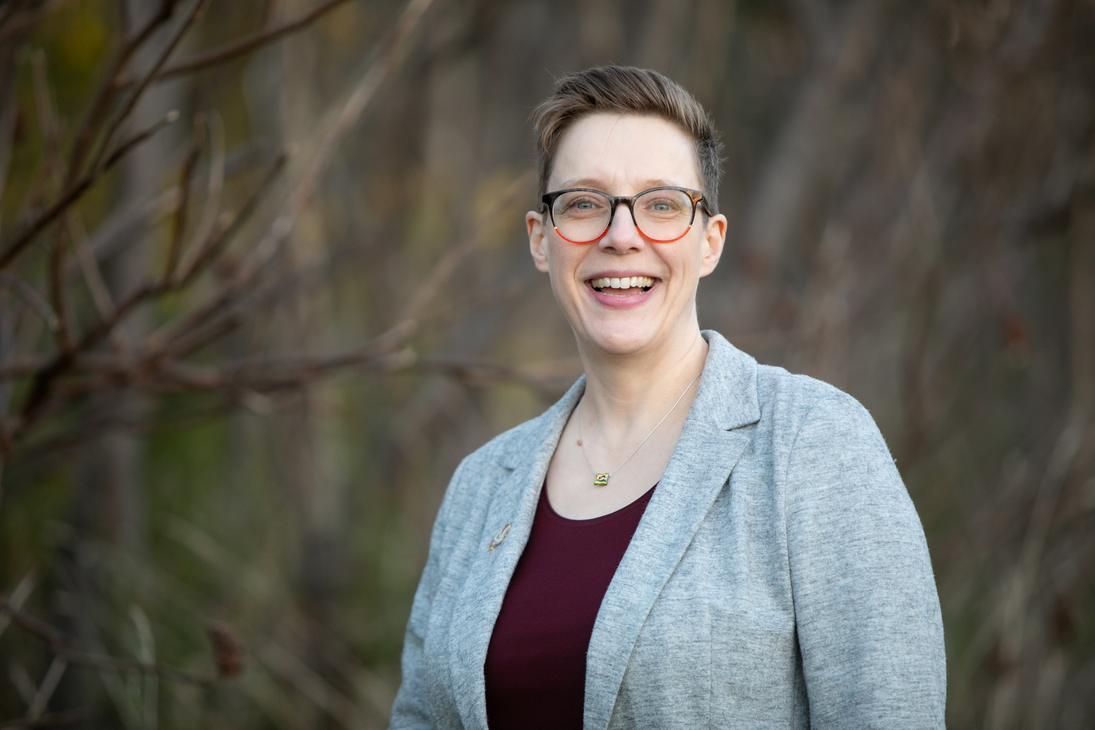 Dr. Anderson, a white woman with short blond hair, blue eyes, and orange glasses, stands smiling in front of some bare branches. She is wearing a maroon top and a grey jacket.