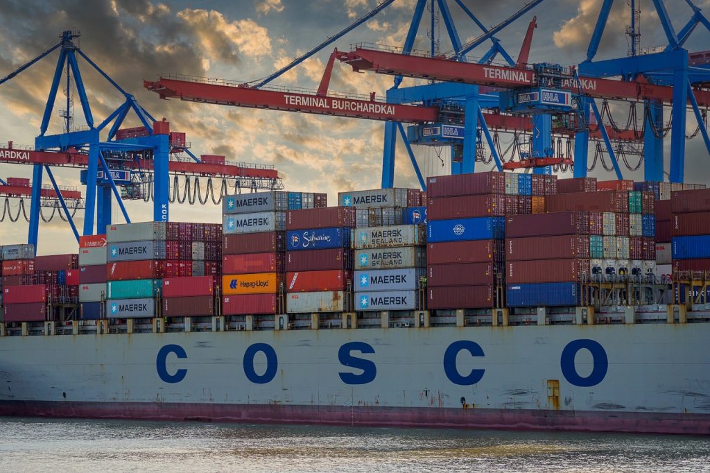 Shipping containers stacked on cargo ship with COSCO written on side; port container cranes in background