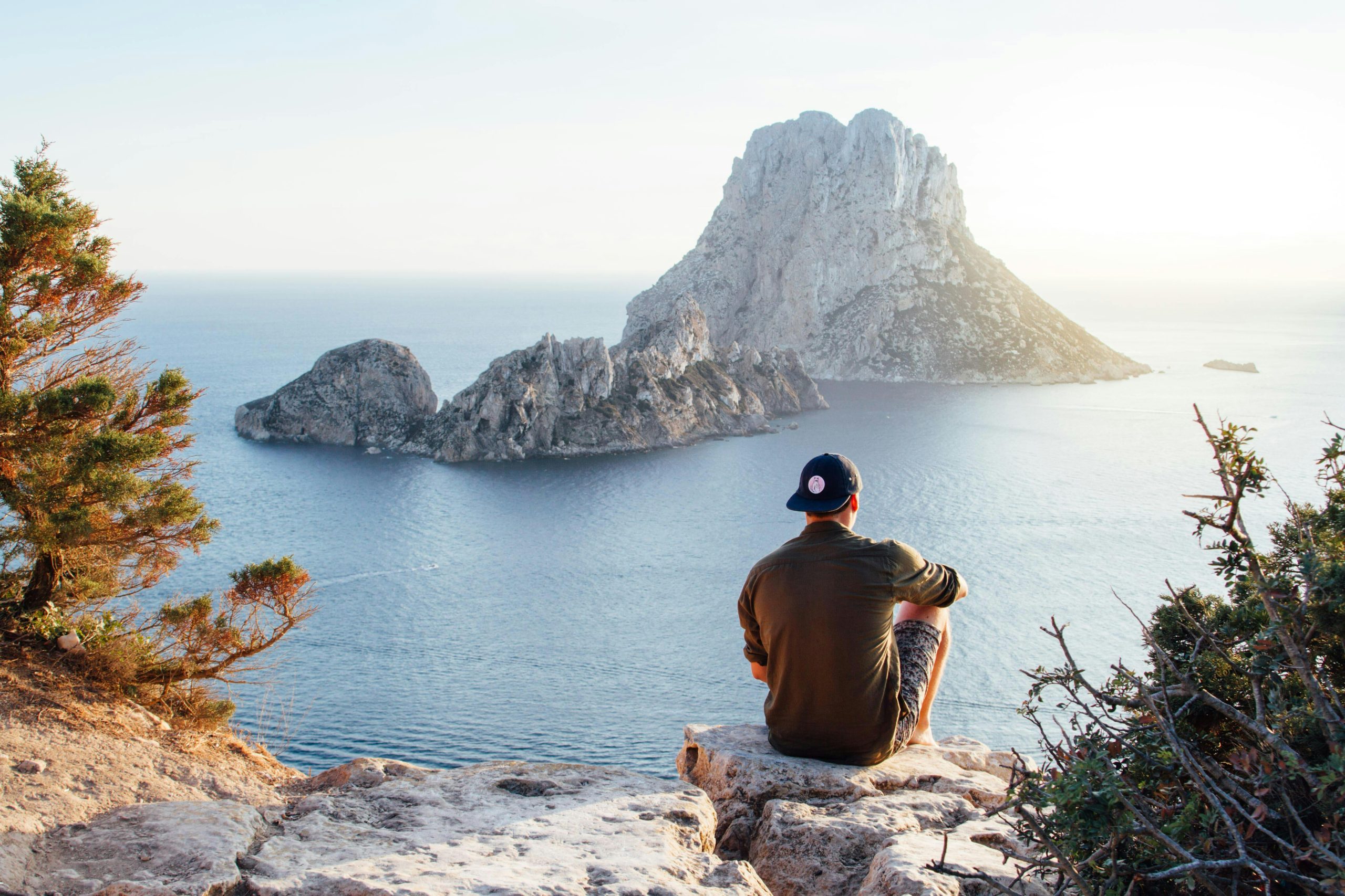 A person in foreground sitting with back to camera on a rock overlooking a lake with large rock formation island in background.