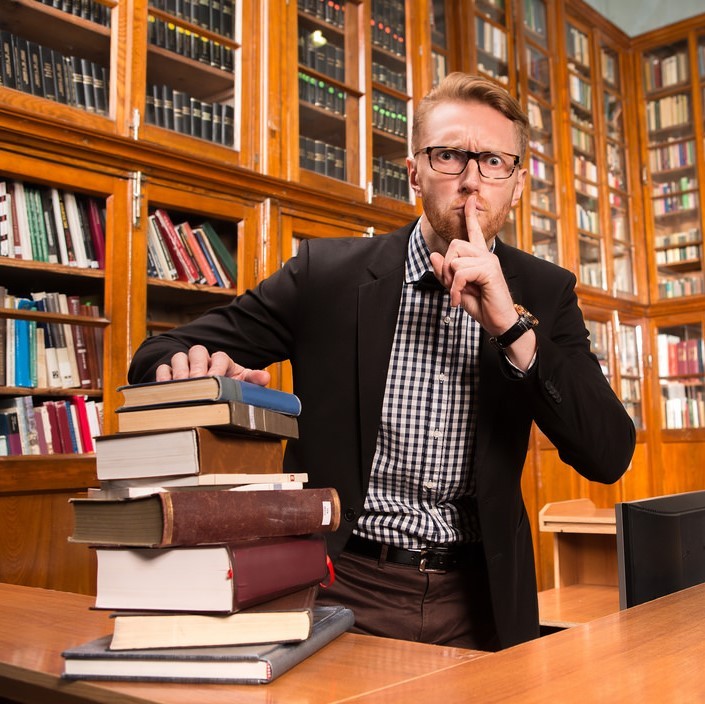 Photo of man sitting at a table in a library with this finger on his lip