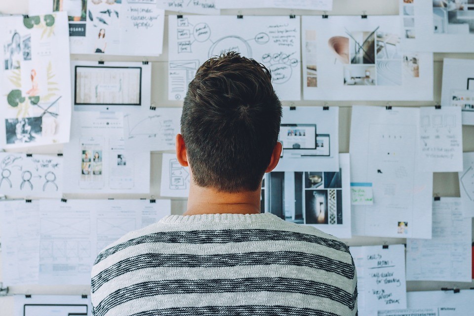 a man looking at a corkboard with pieces of paper, planning and thinking