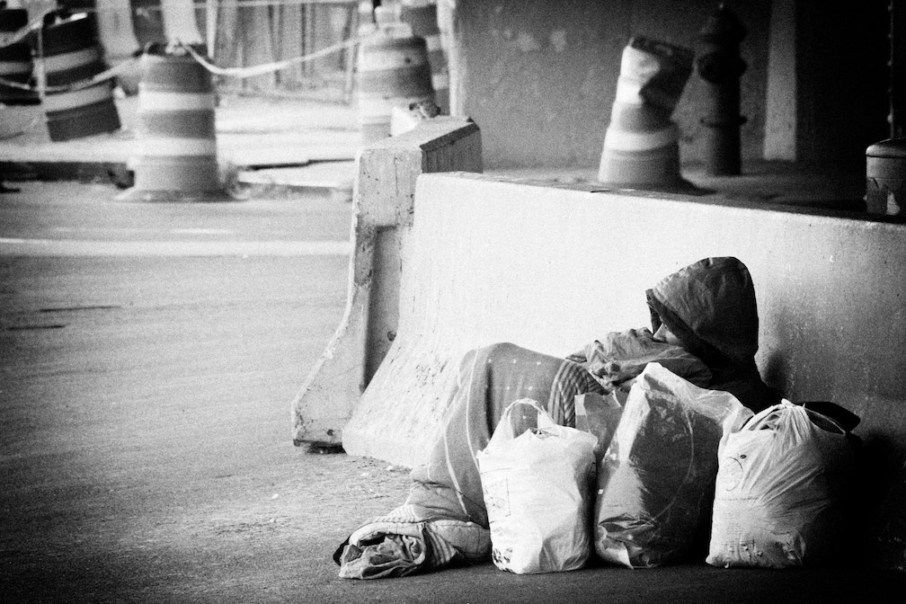 a homeless man lays under a blanket on the street with bags of items around