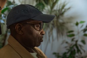 A man wearing a cap and glasses stares thoughtfully off-camera