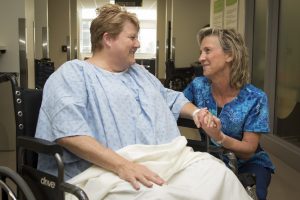 A woman in scrubs kneels next to a patient in a wheelchair to chat.