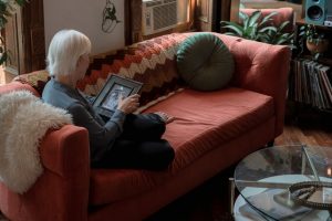 An elderly woman sits on her couch and looks at a framed photo of a man.