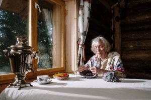 A senior woman knits while seated at a table by a window.